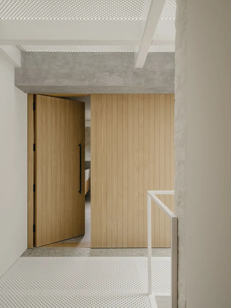Timber sliding door and slatted wall panel beneath a perforated concrete ceiling beam with dappled light