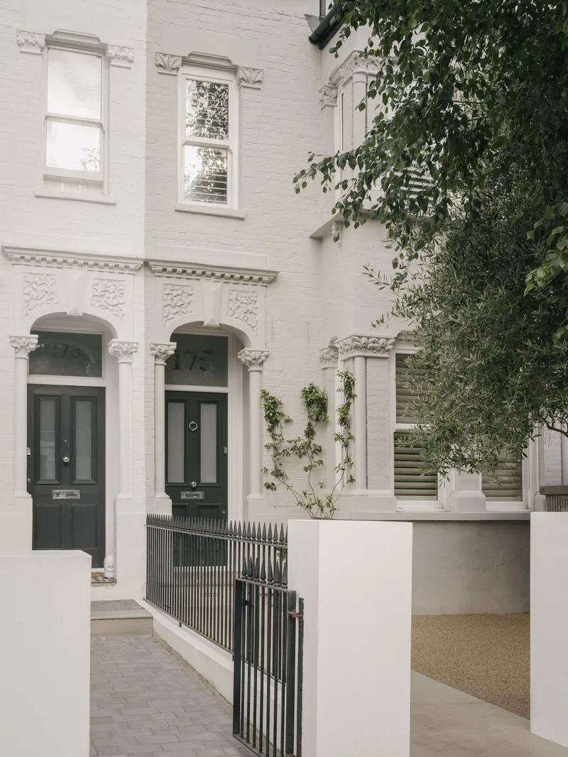 White painted brick facade with arched doorways and climbing vines beside metal railings and gravel path