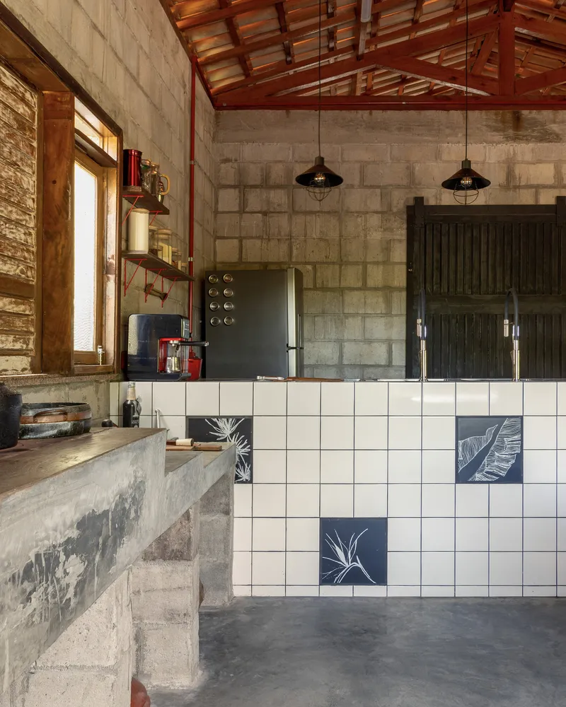 Kitchen with white tile counter featuring botanical motifs beneath exposed timber rafters and concrete block walls