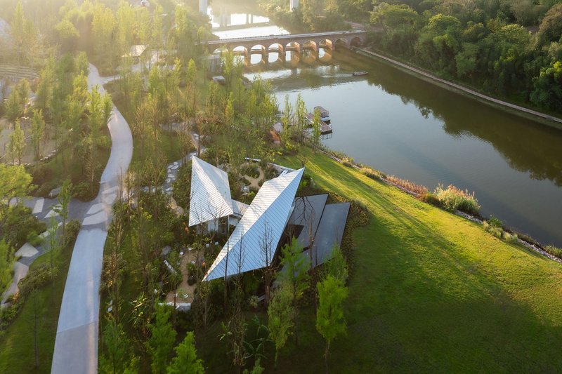 Aerial view of angular metal roof canopies above planted courtyards beside a tranquil pond at sunrise