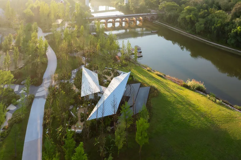 Aerial view of angular metal roof canopies above planted courtyards beside a tranquil pond at sunrise