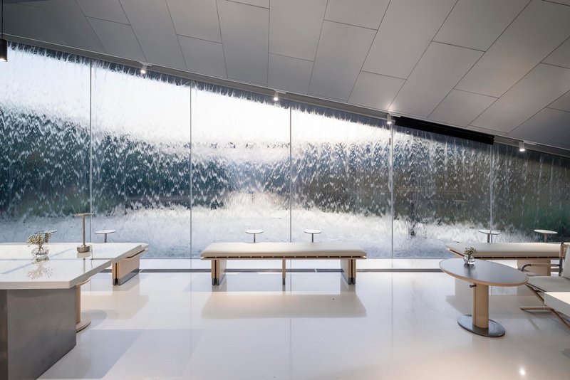 Interior space with continuous glass wall revealing cascading water feature behind wooden benches and tea preparation stations