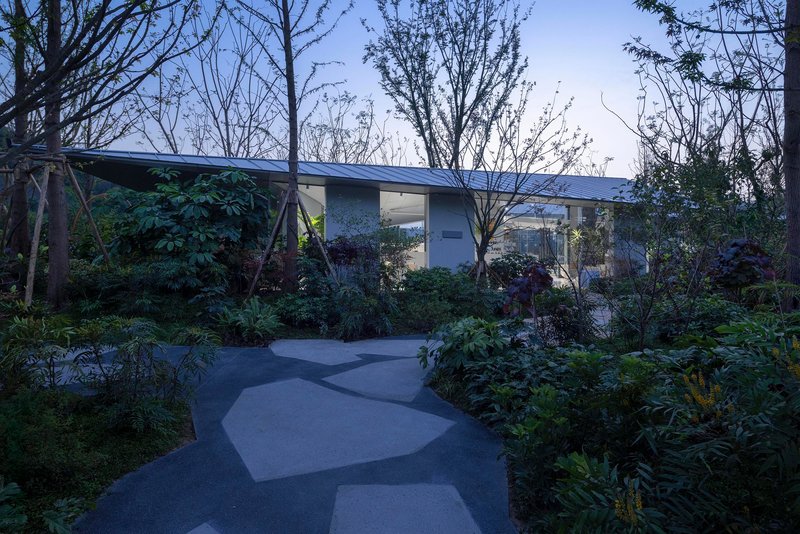 Curving pathway through planted garden leading to low-slung building with folded roof at dusk