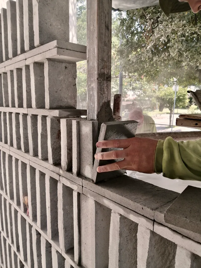 Hand placing custom concrete block into perforated masonry wall during construction with trees visible beyond