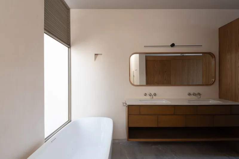 Bathroom with freestanding tub, timber vanity unit and rounded mirror beneath a lowered roman shade