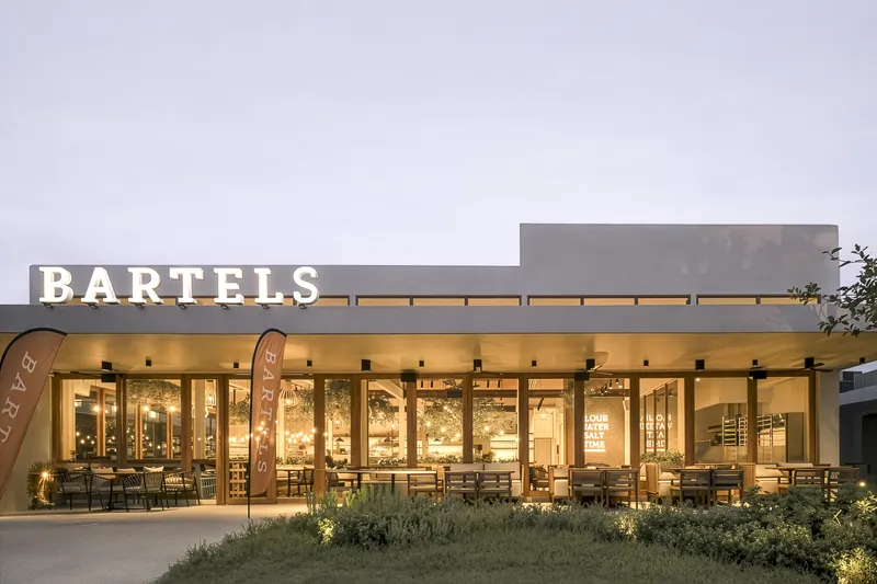 Glass-walled restaurant facade with interior lighting glowing through planted forecourt at twilight