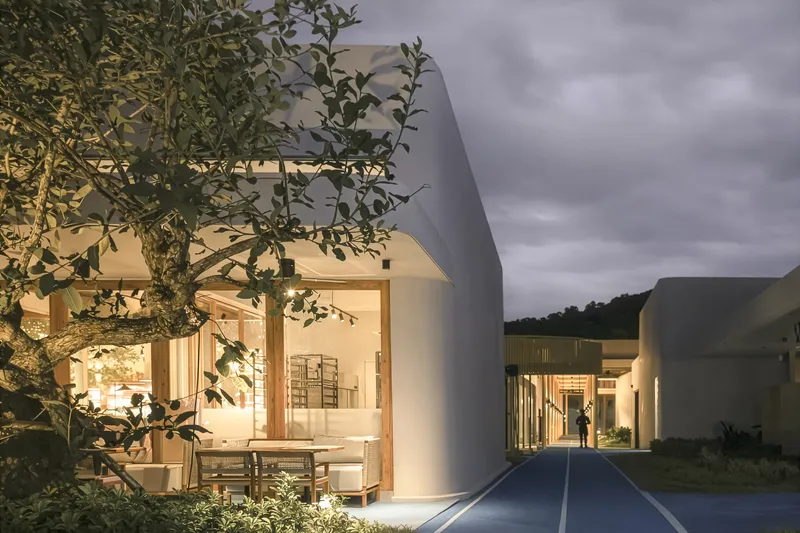 Illuminated white volumes with glazed openings framed by a tree at dusk
