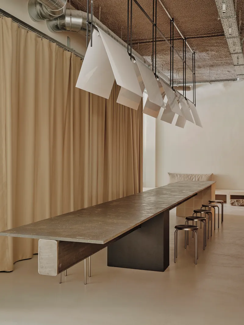 Long communal table with steel stools beneath a row of suspended white fabric pendants and exposed cork ceiling