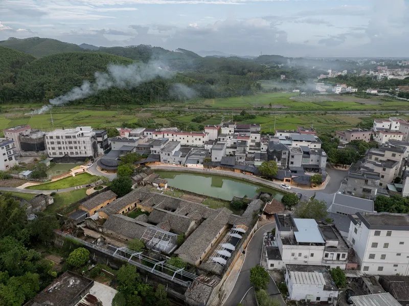 Aerial view of a village with tiled roofs surrounding a central pond and green fields in mist