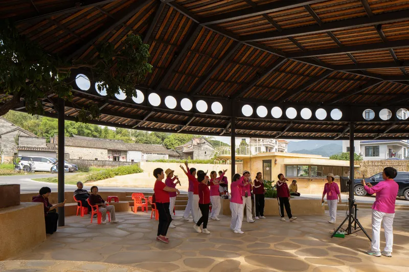 Timber pavilion with exposed bamboo rafters and circular window openings providing shelter for a group in matching attire