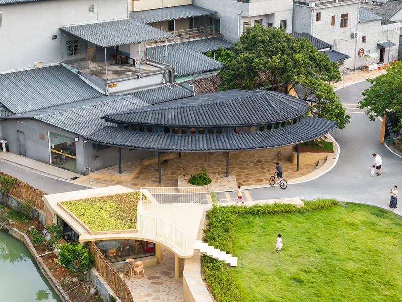 Aerial view of a curved tile roof pavilion with circular openings adjacent to a green roof and residential buildings