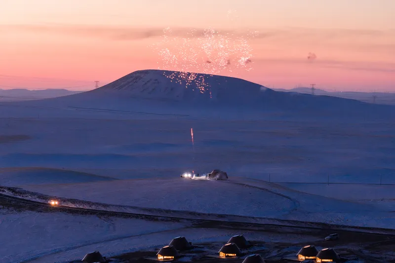 Cluster of dome-shaped shelters in a snowy field at dusk with illuminated volcano erupting in distance