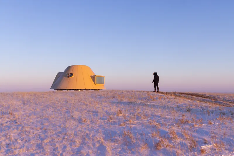 Person standing beside dome-shaped shelter on a snow-covered ridge in golden sunset light