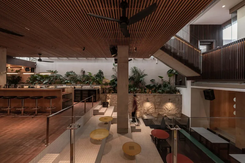 Double-height interior with timber-slatted ceiling, planted planter wall, and circular stools around glass-railed mezzanine