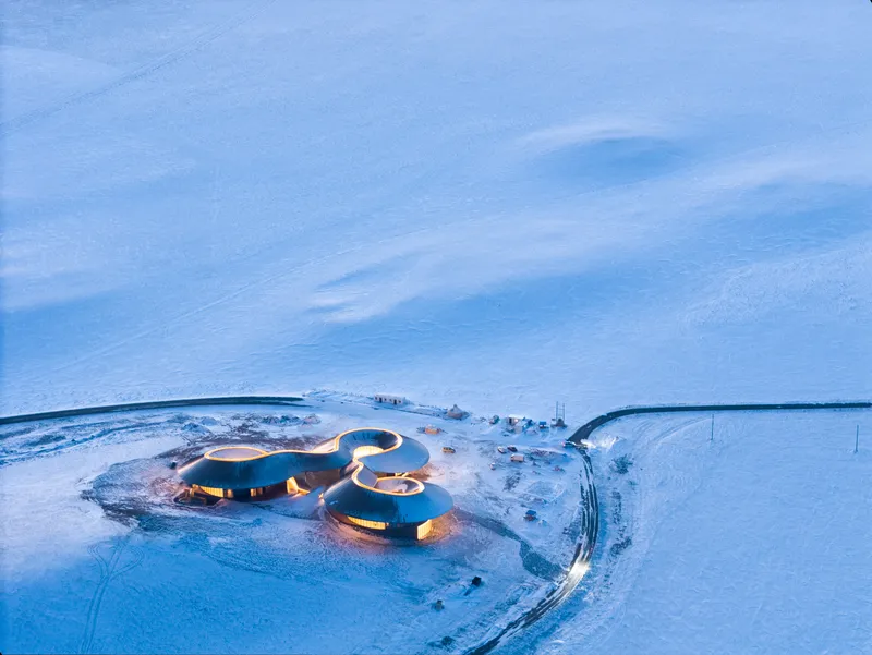 Dusk aerial with the building's interior glowing warm against the blue snow