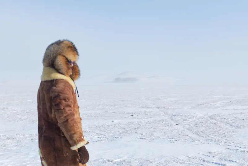 A figure in a fur-trimmed shearling coat looking across the snow steppe