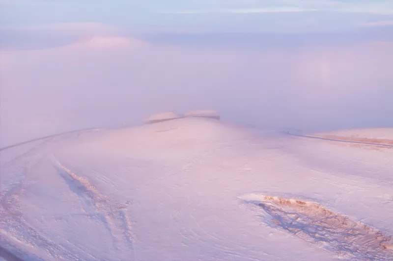 Distant view of the building tucked into a snowy ridge under pink sunset clouds