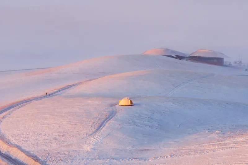 A small lit yurt-like structure in the snow with the visitor center on the horizon