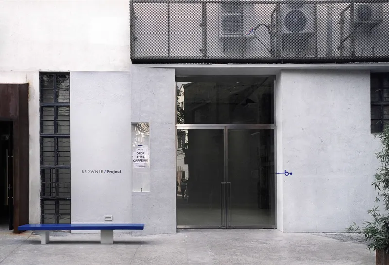 Street facade with glass entrance doors and perforated metal balcony above a blue painted bench