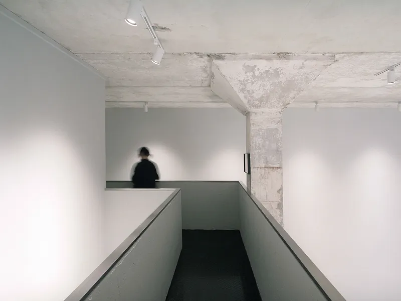 Ramp descending between white walls with concrete ceiling and figure visible on mezzanine beyond