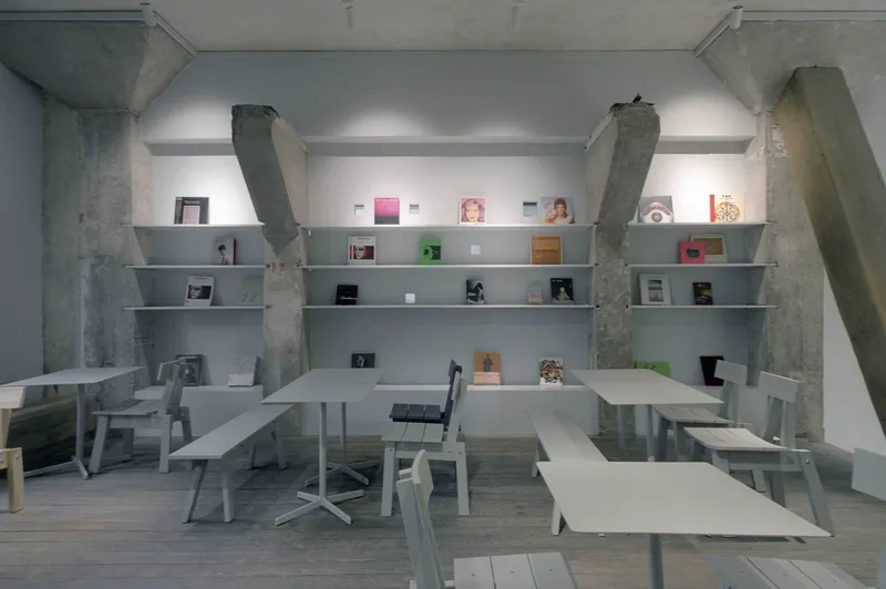 Interior workspace with white metal tables and benches beneath wall-mounted shelving and exposed concrete columns