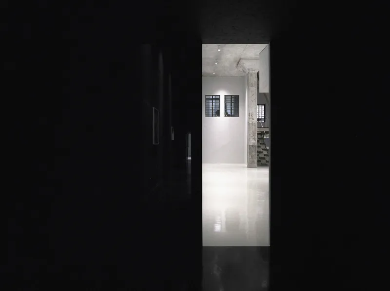 View through a dark corridor into a white-walled gallery space with concrete column and ceiling spotlights