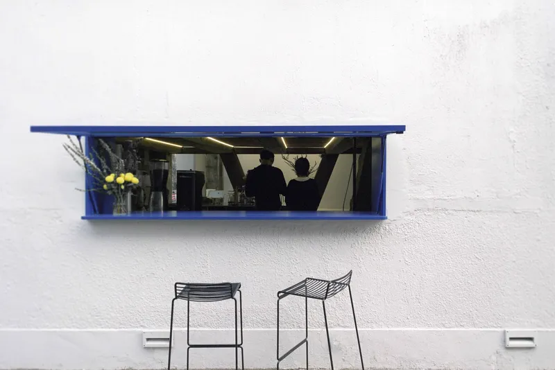 Blue-framed service window with mirrored interior set into white exterior wall above two wire stools