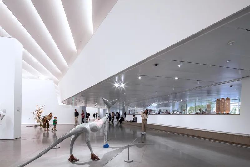 Gallery space with sculptural figures beneath a hovering metallic ceiling plane and skylights