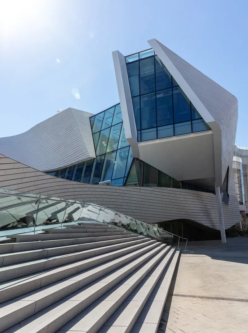 Upward view of the ribbed metal facade intersected by angled glass curtain walls under clear blue sky