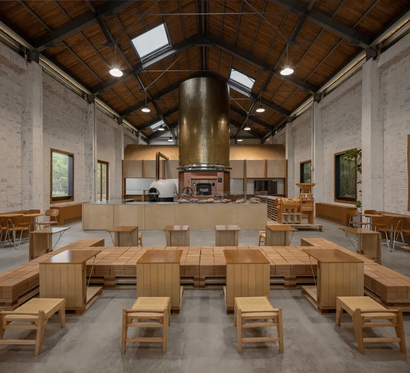 Interior dining hall with timber ceiling, exposed steel trusses, central bronze chimney and wooden communal tables
