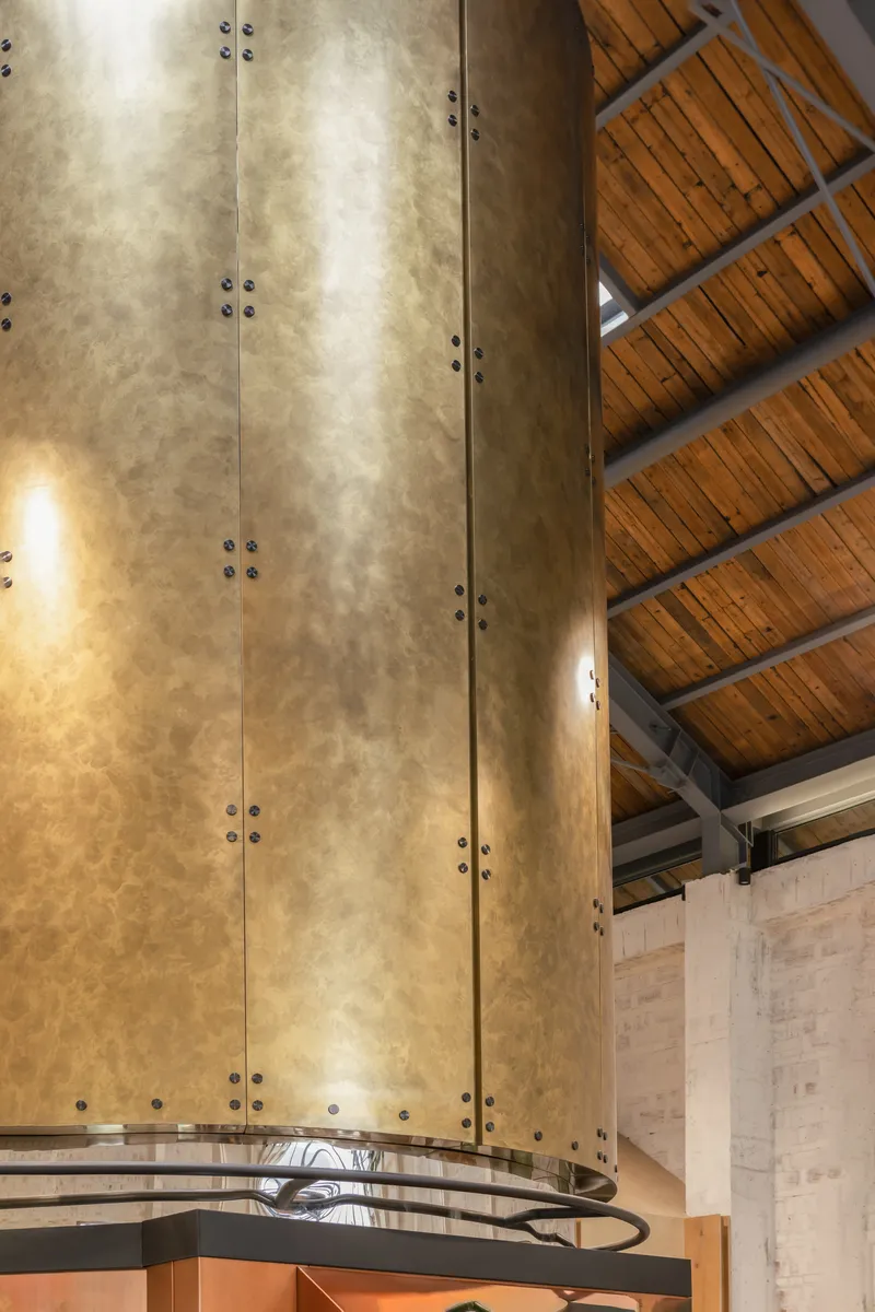 Brass-clad chimney flue with visible bolts rising through timber-lined ceiling with exposed steel beams