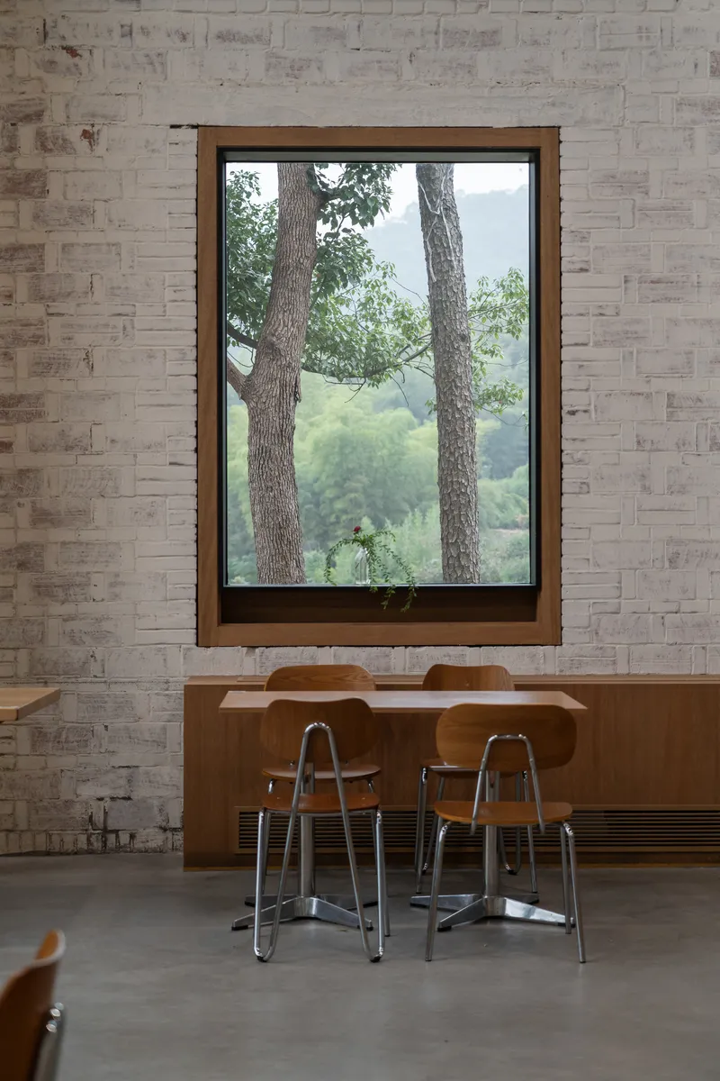 Timber-framed window set into a textured pale brick wall above a wooden bench with metal stools