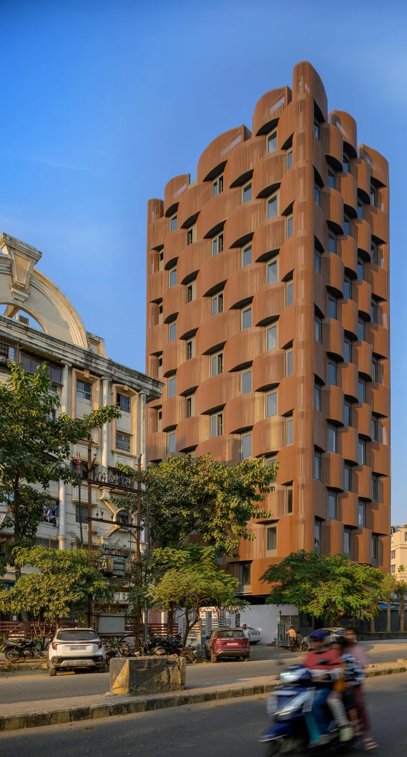 Street view of the tower with undulating terracotta brise-soleil panels beside a tree-lined road with passing motorcycles