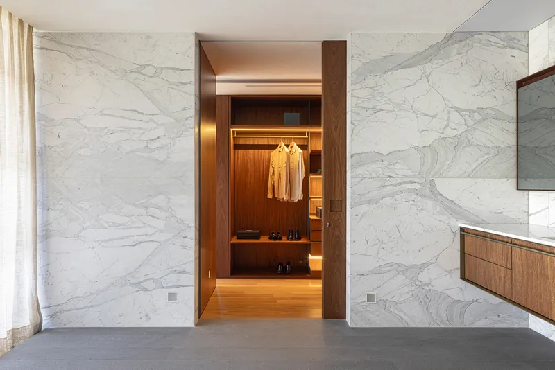 White marble wall with timber doorway opening into a backlit wardrobe with hanging garments