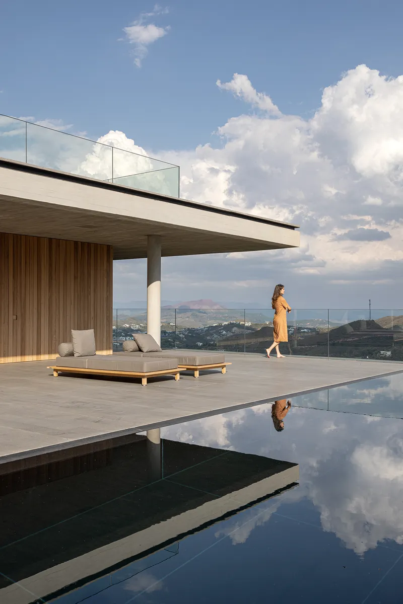 Infinity pool deck with a figure walking past timber clad pavilion under billowing afternoon clouds
