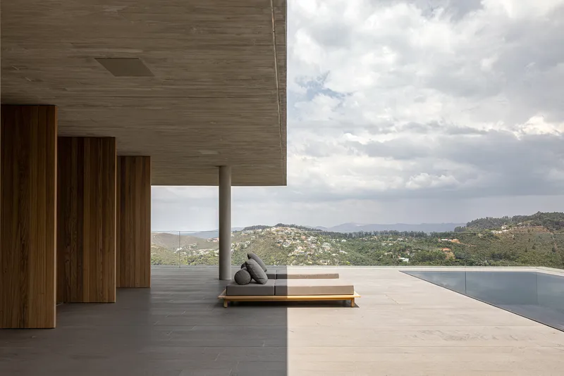 Covered terrace with infinity pool and low daybed facing hillside settlement under cloudy sky