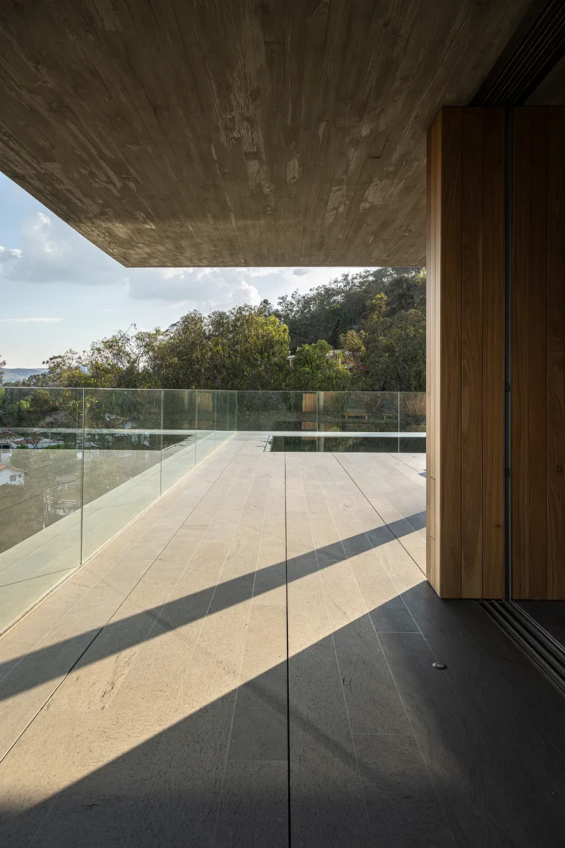 Glass-walled corridor with timber panels and stone flooring casting afternoon shadows across the terrace