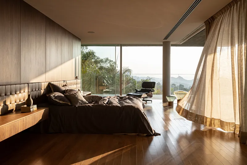 Bedroom with angled window wall and sheer curtains overlooking mountain ridges in morning light