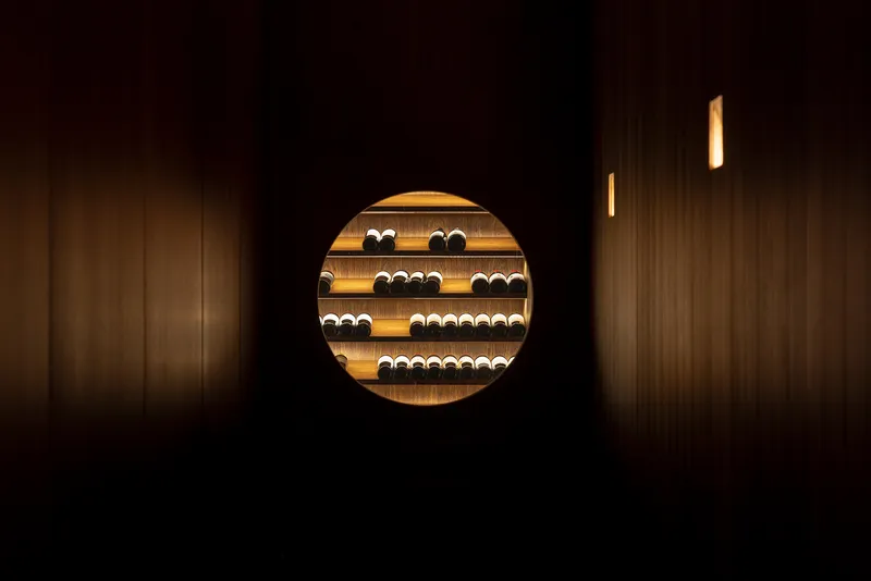 Circular aperture in a dark timber wall framing illuminated wine storage shelves