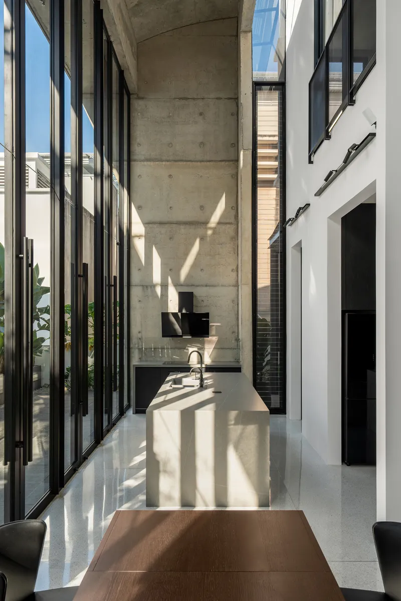 Long view through kitchen corridor with vertical black steel mullions casting geometric shadows on white terrazzo counter