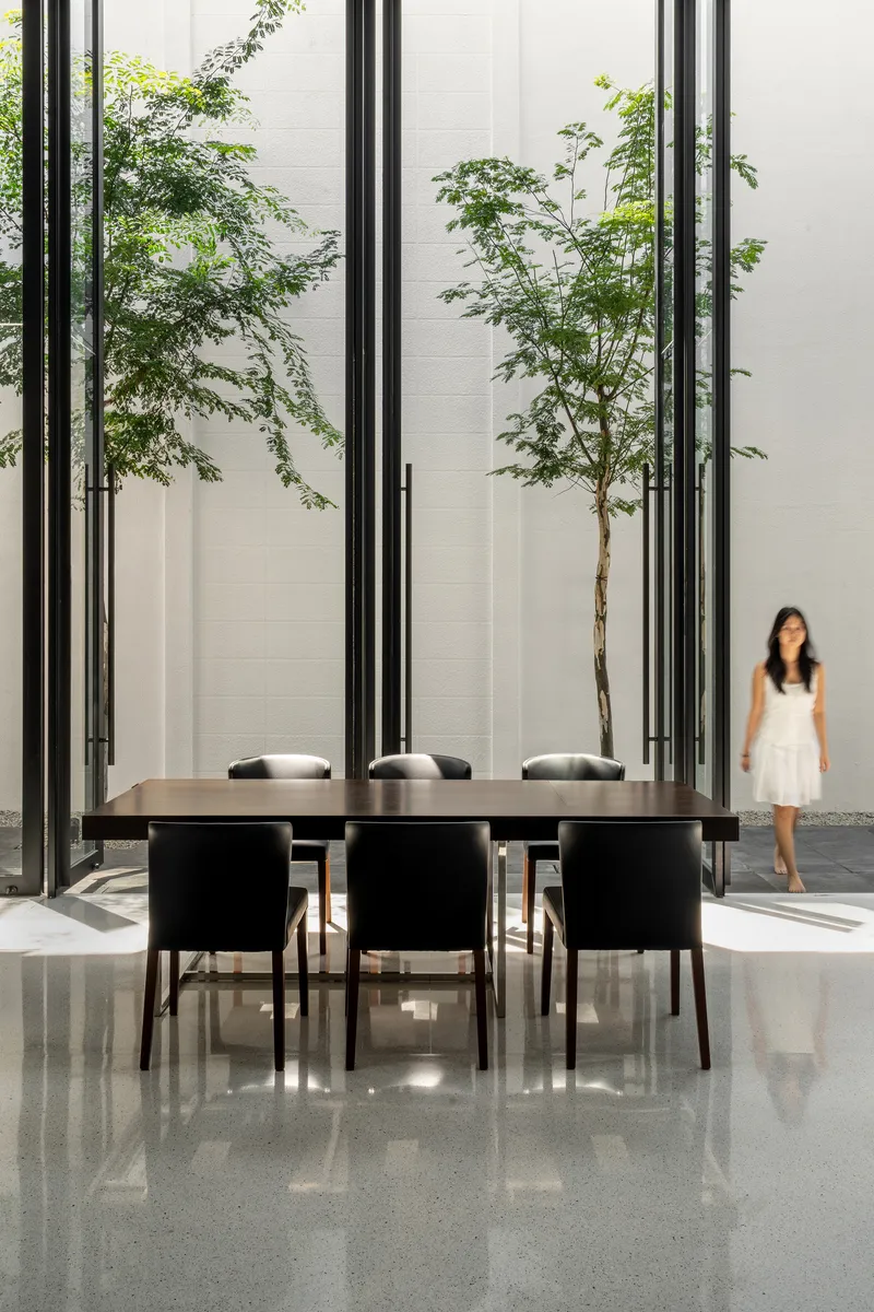 Dining table surrounded by steel frame screens with young trees and a figure in the courtyard