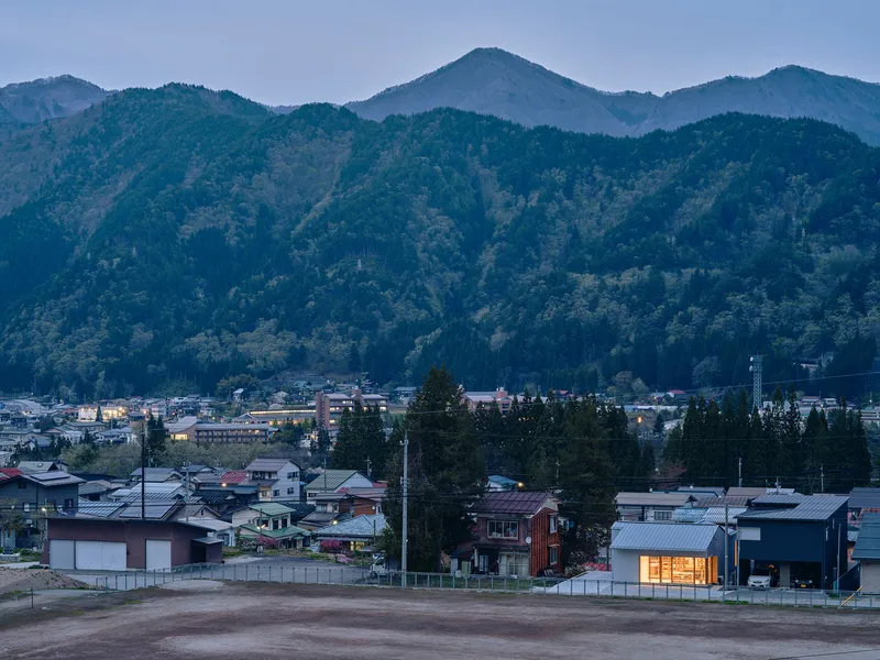 Illuminated single-story house with timber slatted facade at dusk in a village below forested mountains