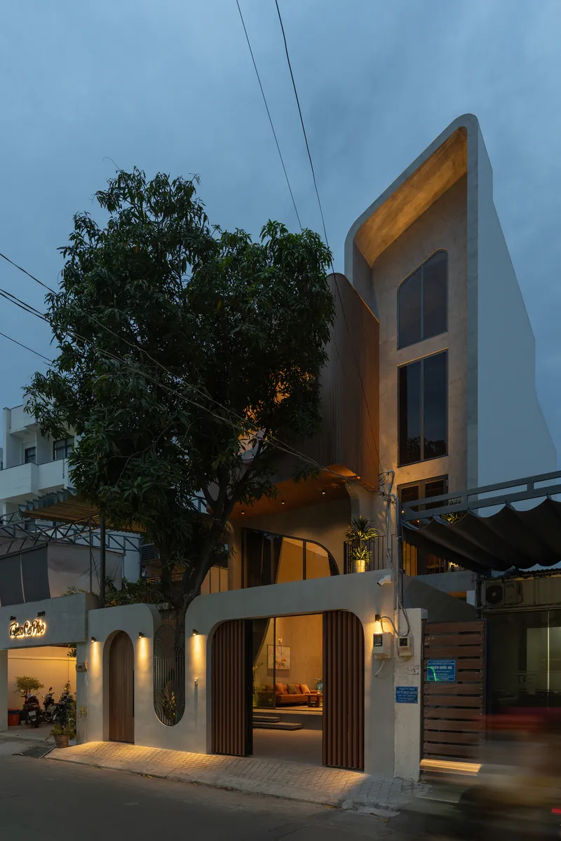 Street facade with arched openings and vertical timber slats illuminated at dusk beside a large tree