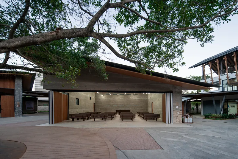 Open-air assembly space with rows of benches under a cantilevered roof framed by mature tree branches