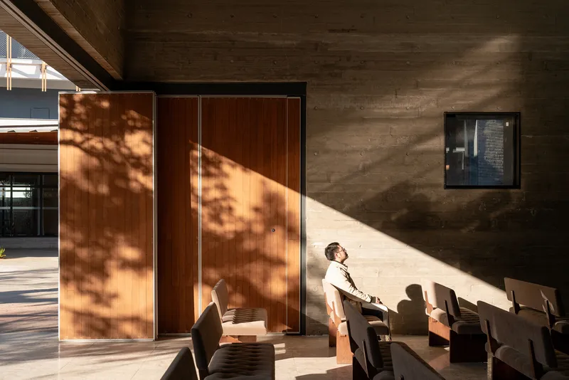 Interior seating area with board-formed concrete wall receiving sharp diagonal sunlight and tree shadows through clerestory