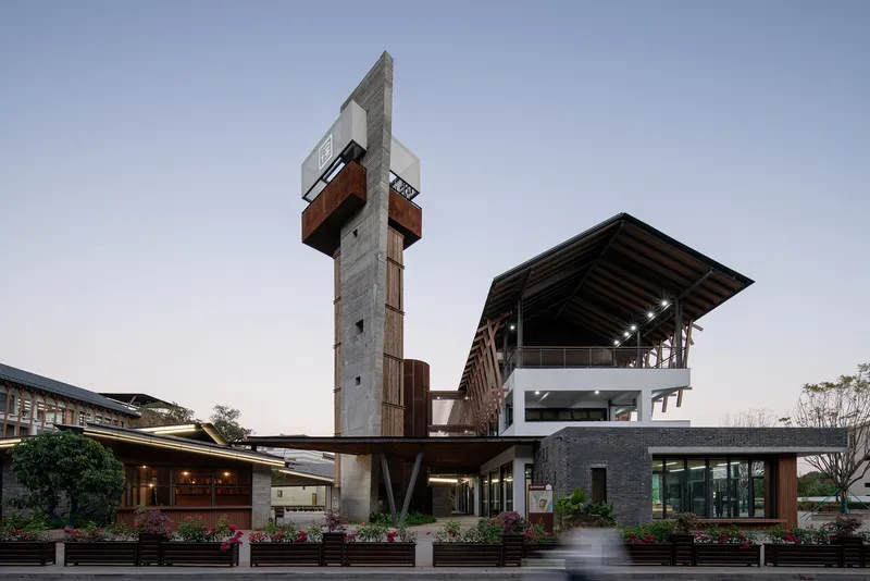 Concrete tower with cantilevered weathered steel volumes beside low-rise pavilions at dusk
