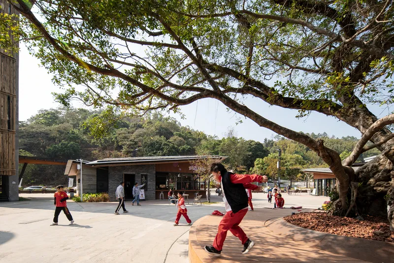 Open courtyard with low-rise pavilions and children playing beneath a large spreading tree