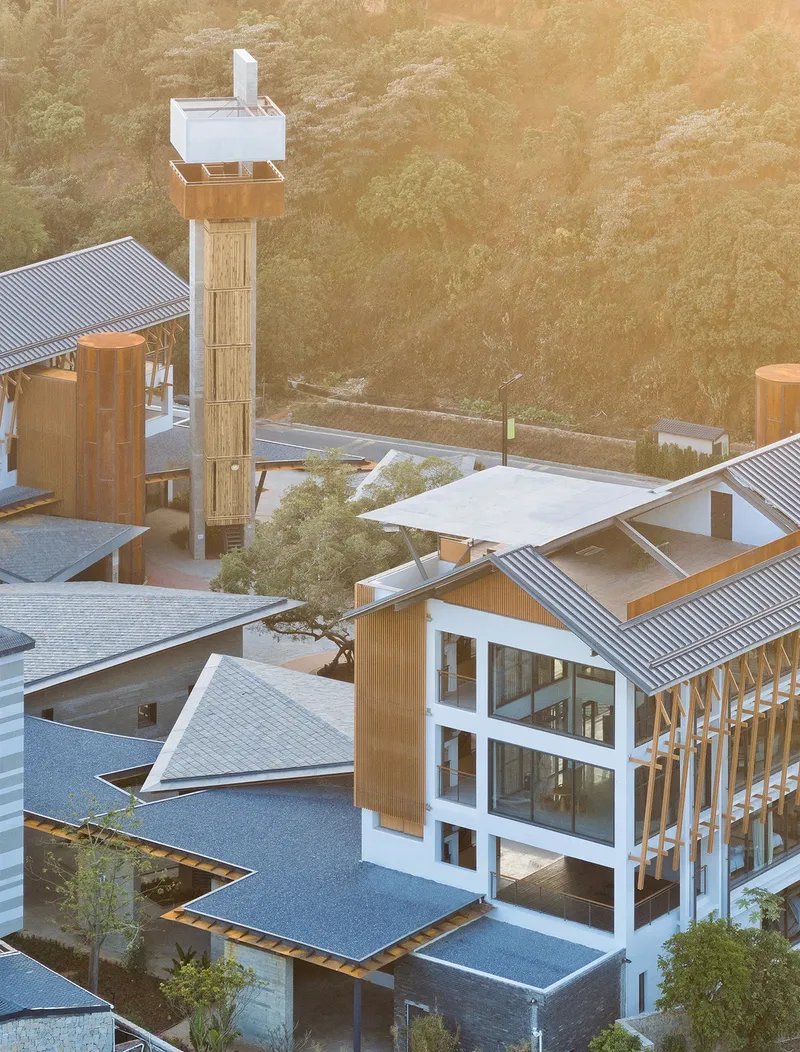 Aerial view of clustered volumes with corrugated metal roofs and timber-clad tower at golden hour