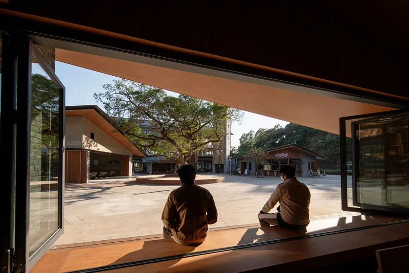 View from interior timber bench through open corner glazing where two seated figures look toward courtyard at dusk