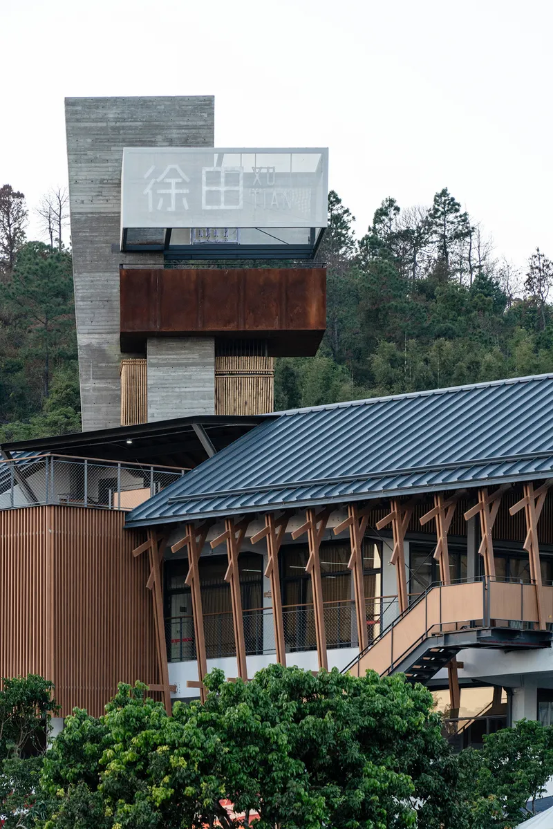Tower with stacked concrete and timber volumes rising above a corrugated metal roof with forested hillside